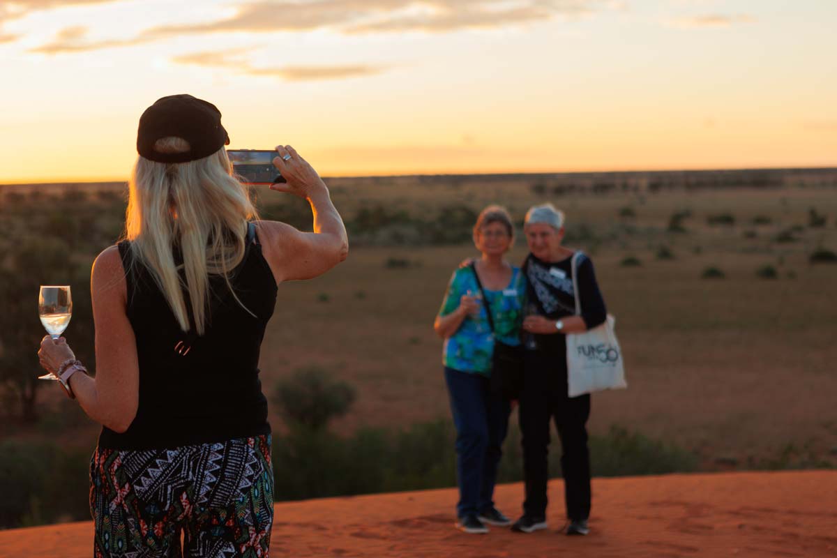 Woman holding glass of wine takes photo of two ladies on sandhill with sunset